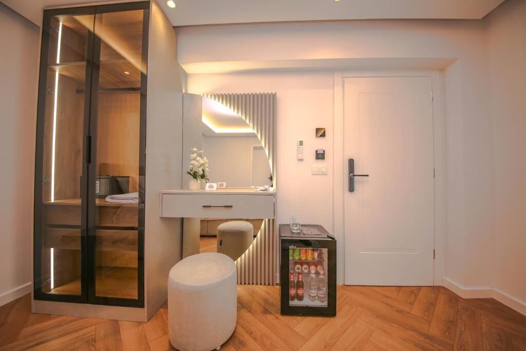 A modern hotel room with a wooden floor, a glass-enclosed wardrobe, a white vanity with a mirror, a small ottoman, and a minibar.