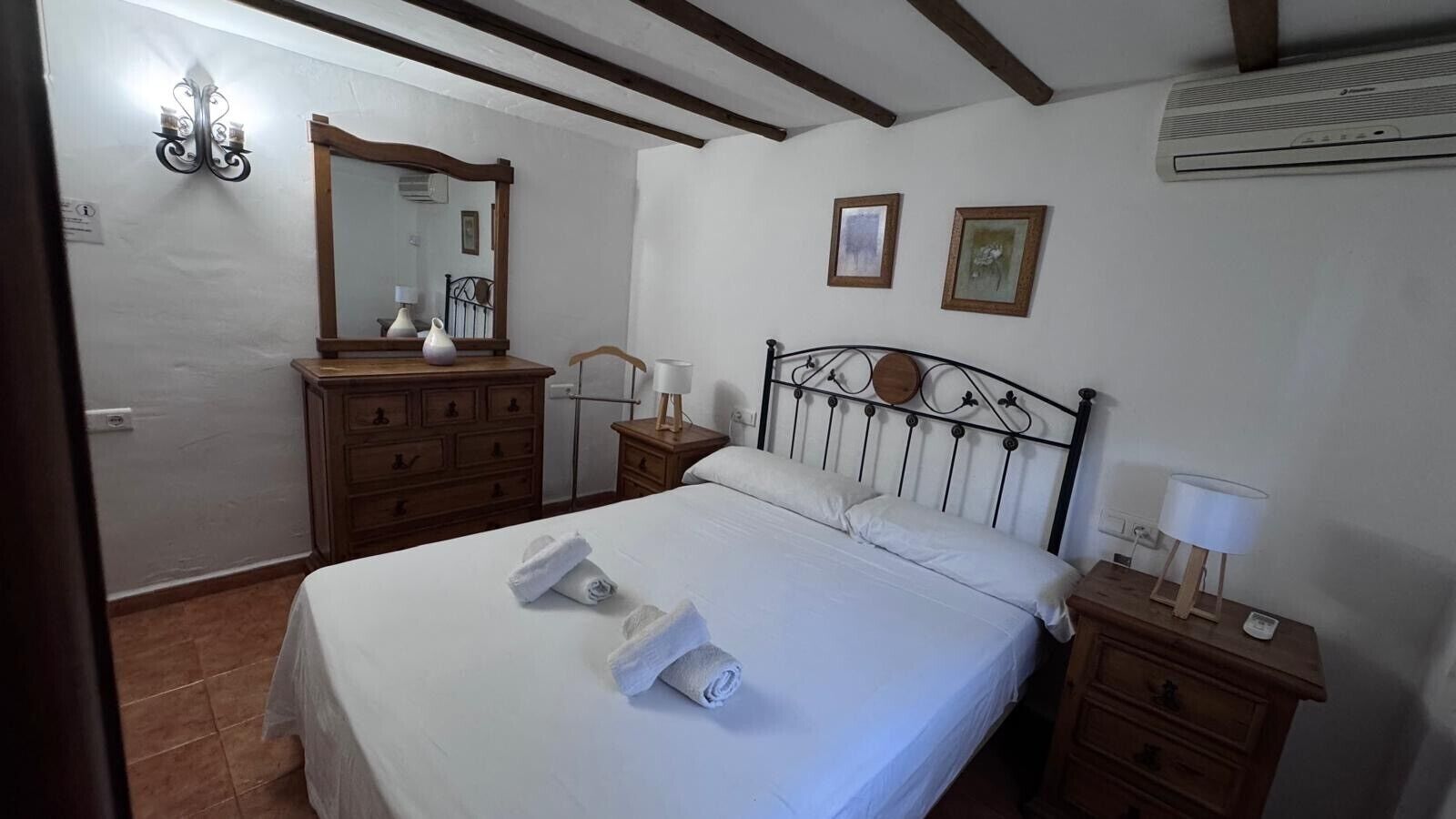 A bedroom with a bed, wooden dresser, bedside tables, a mirror, and two framed pictures on the wall.