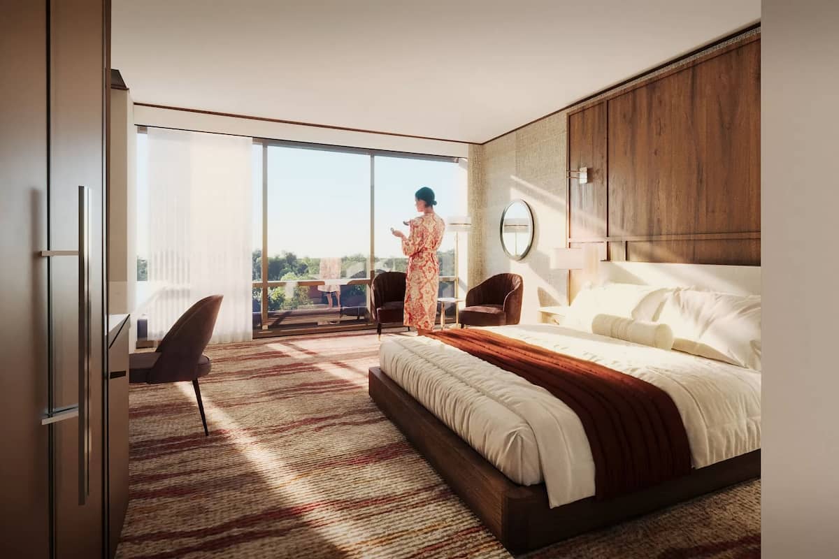 A modern hotel room with a large bed, a sitting area with a chair, a balcony with a view, and a large window.