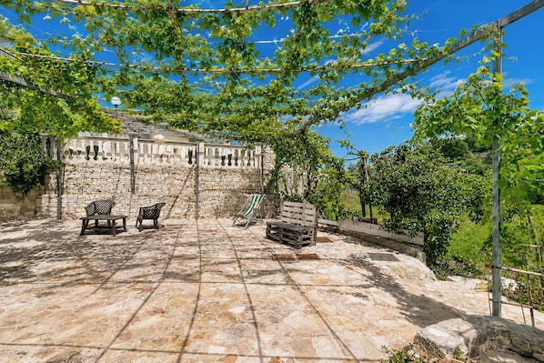 A stone patio with outdoor furniture, a stone wall, and a pergola with climbing plants.