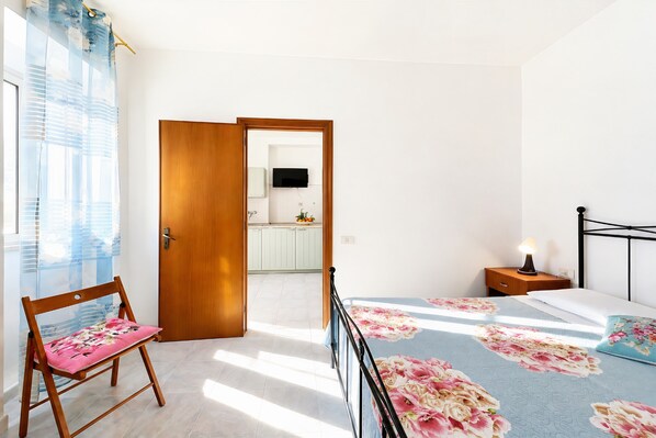A bedroom with a bed, a nightstand, a chair, a wooden door, and a view into the kitchen.