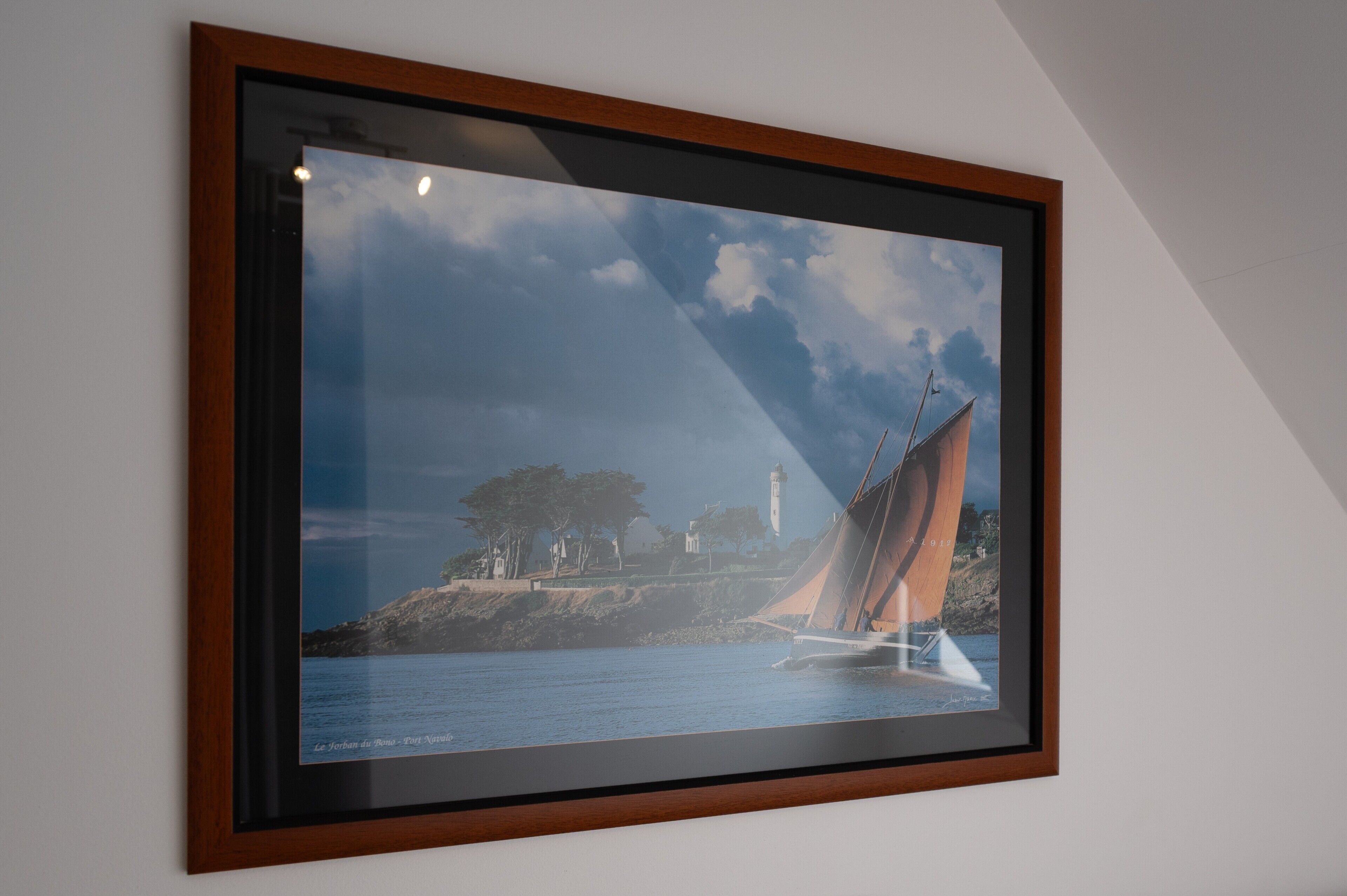 A framed photograph of a sailboat on water with a lighthouse and trees in the background.