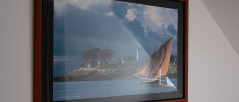 A framed photograph of a sailboat on water with a lighthouse and trees in the background.