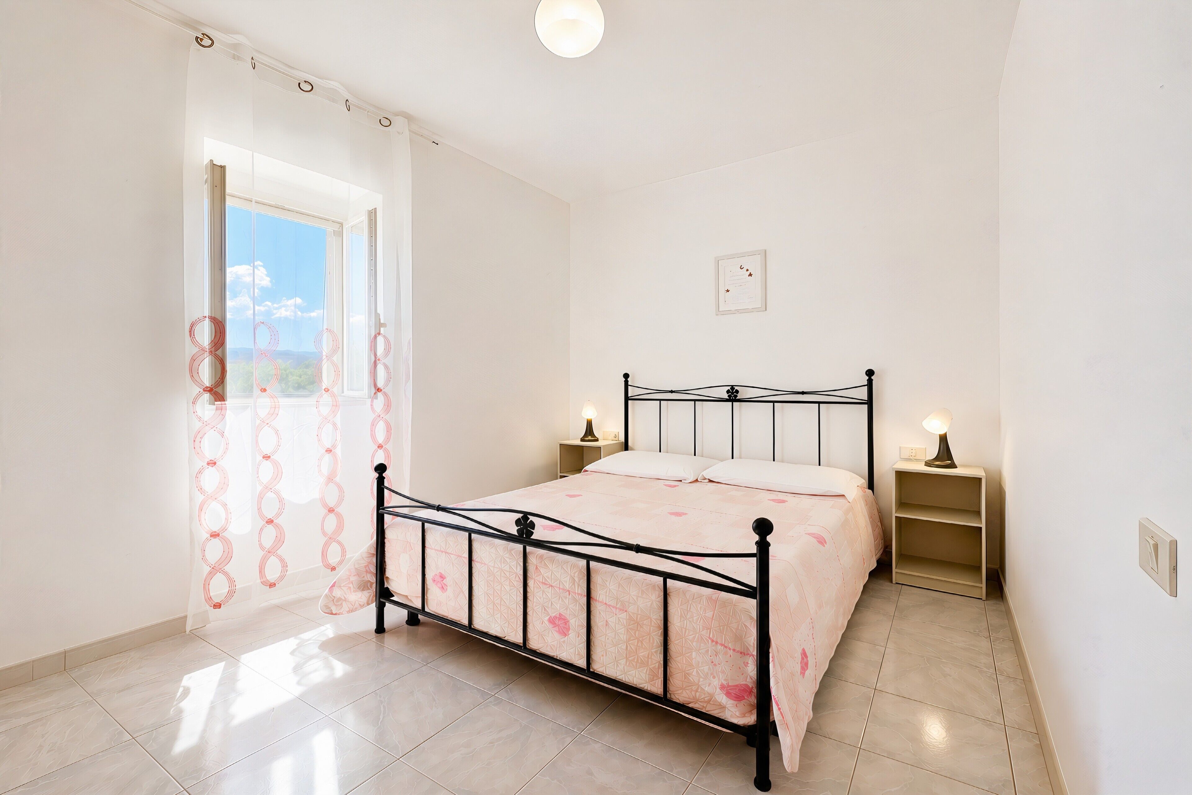 Un dormitorio con una cama, dos mesitas de noche, una ventana con cortinas e iluminación en la pared.