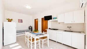 Uma cozinha compacta com armários brancos, uma mesa de jantar com cadeiras, um frigorífico pequeno e uma TV fixada acima do balcão.