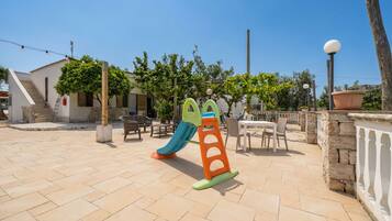 Un parque infantil con tobogán y estructura para escalar, mobiliario exterior y un edificio con escaleras.