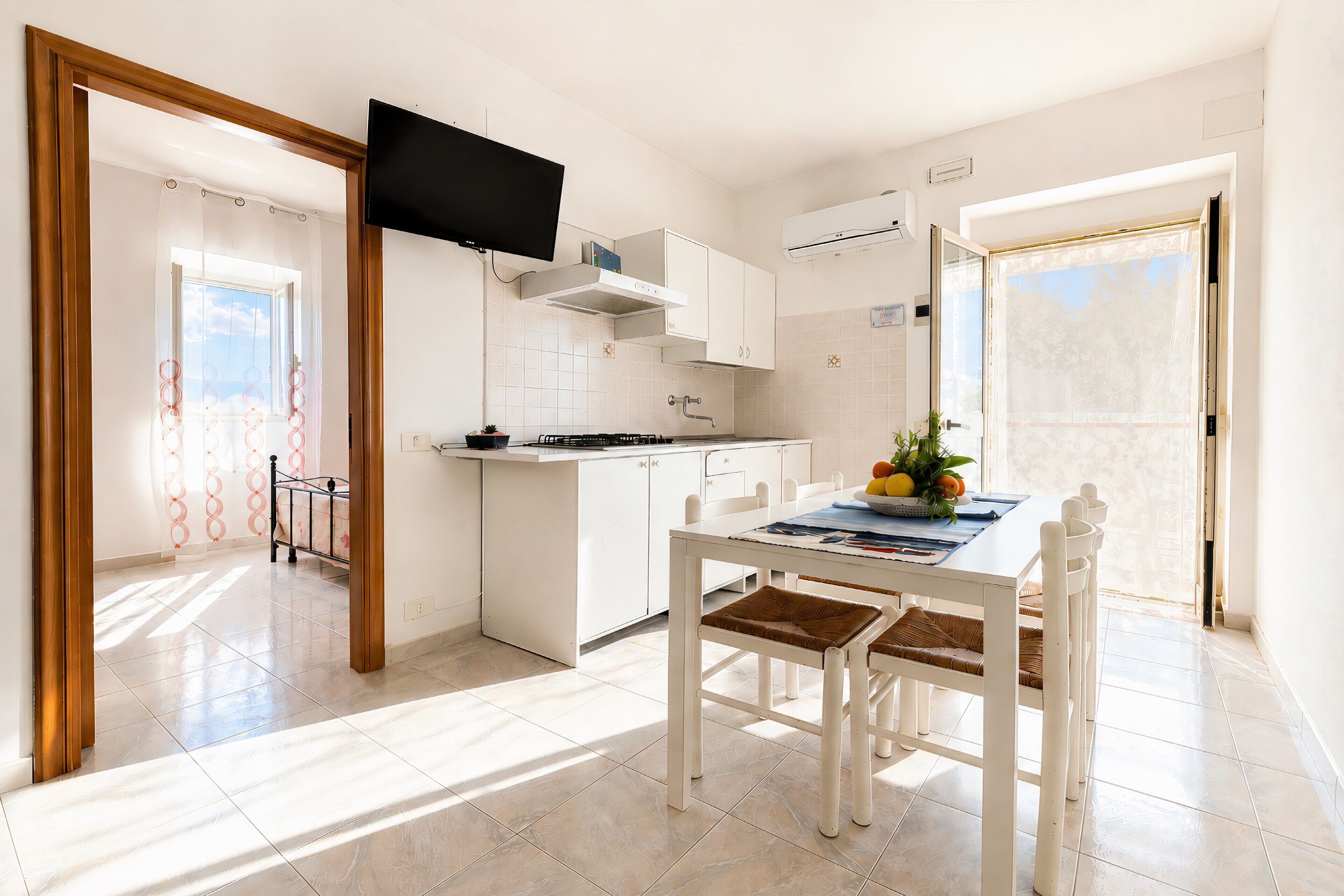 Una cocina moderna con armarios blancos, una mesa de comedor con sillas, un televisor de pantalla plana sobre la encimera y vistas al exterior a través de una puerta.