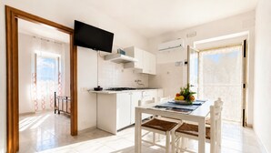 Uma cozinha moderna com armários brancos, uma mesa de jantar com cadeiras, uma televisão de tela plana instalada acima do balcão e vista para o exterior através de uma porta.