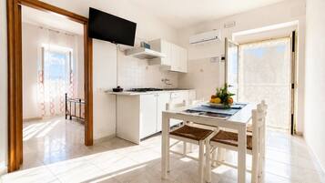 Una cocina moderna con armarios blancos, una mesa de comedor con sillas, un televisor de pantalla plana sobre la encimera y vistas al exterior a través de una puerta.