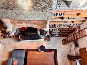 A stone-walled interior with a fireplace, bookshelves, a wooden table, and a staircase.