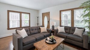 A modern living room with a sectional sofa, a coffee table, and large windows with blinds. - LOCATION! 3BR Near UNMC, Creighton, UNO & Downtown (Omaha)