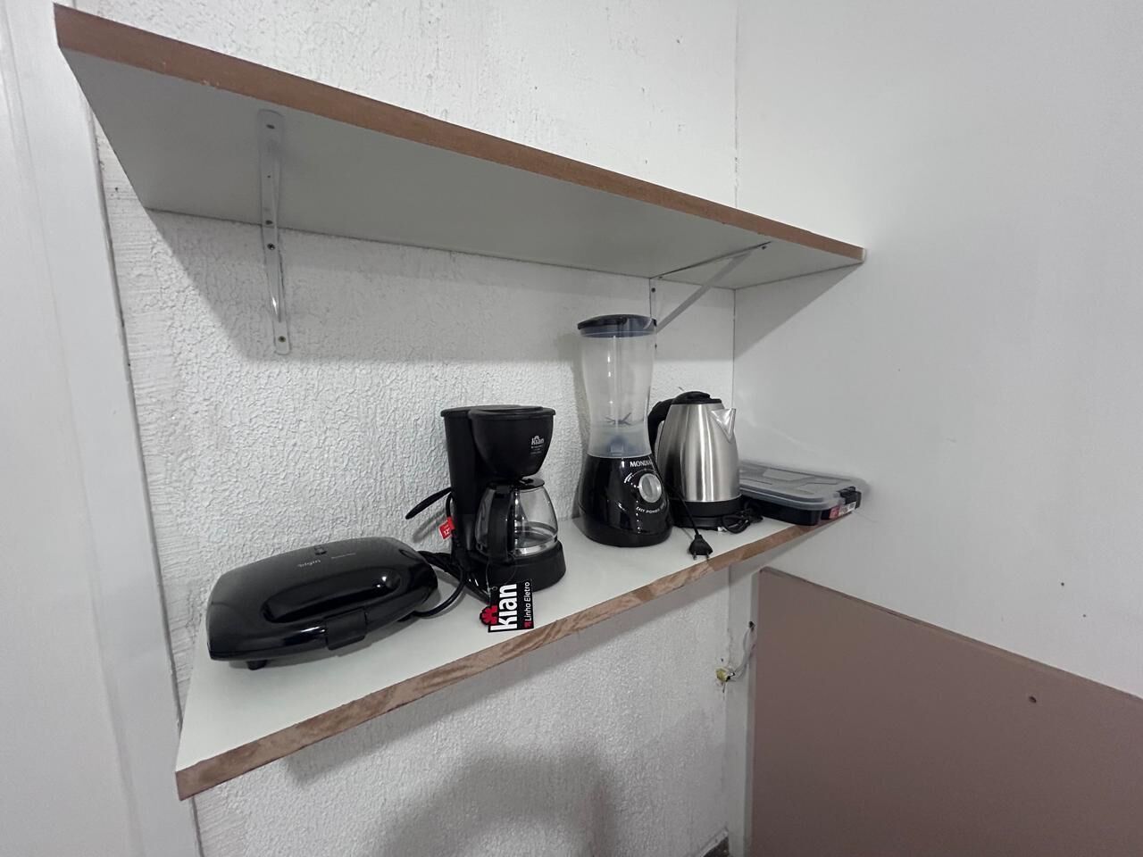 A kitchenette with a coffee maker, blender, kettle, and toaster on a white shelf.