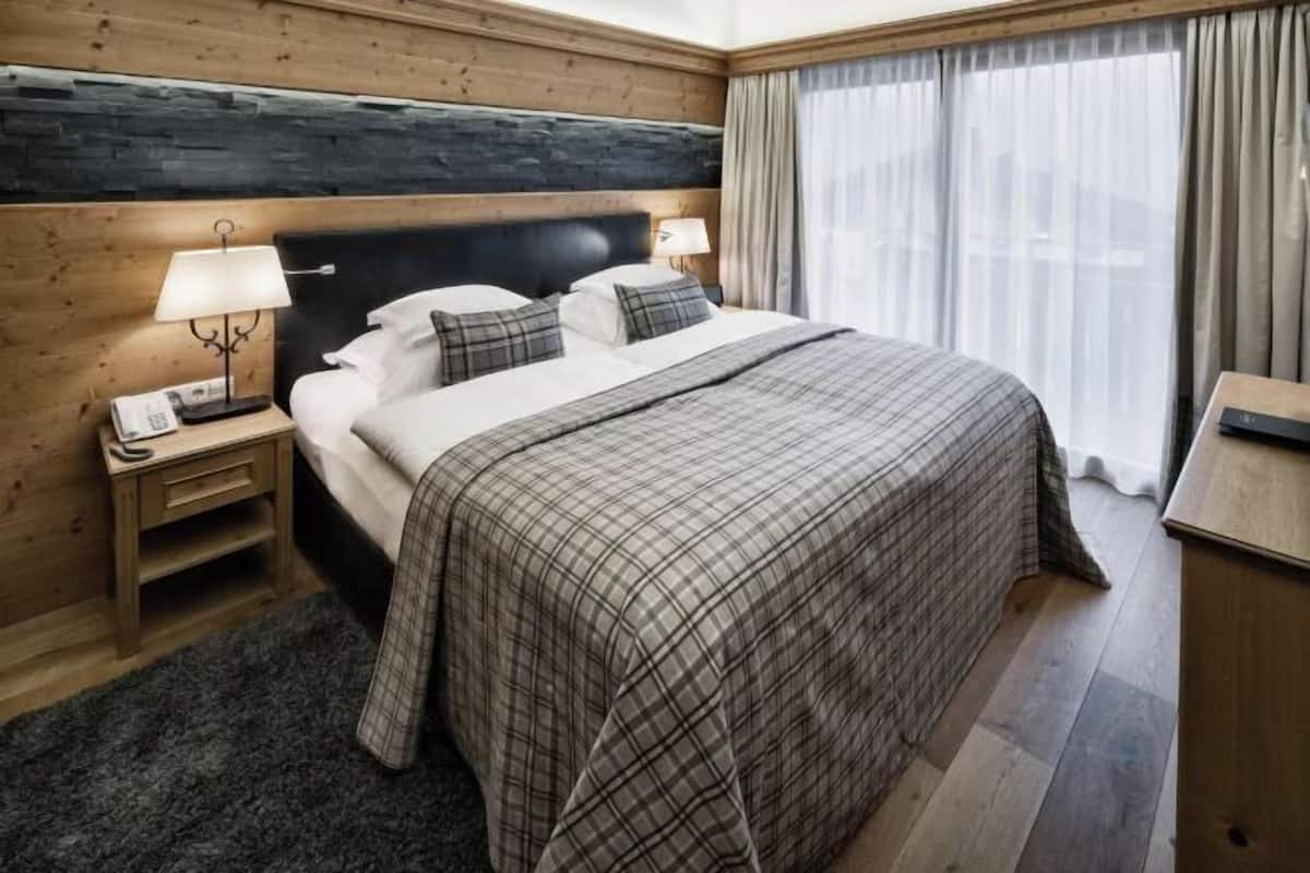 Ein Hotelzimmer mit Bett, Nachttisch mit Lampe, Holz-Kopfteil und einem Fenster mit Vorhängen.