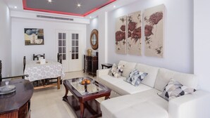 A living room with a white sofa, a coffee table, a dining table with a patterned tablecloth, and artwork on the walls.