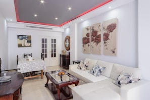 A living room with a white sofa, a coffee table, a dining table with a patterned tablecloth, and artwork on the walls.