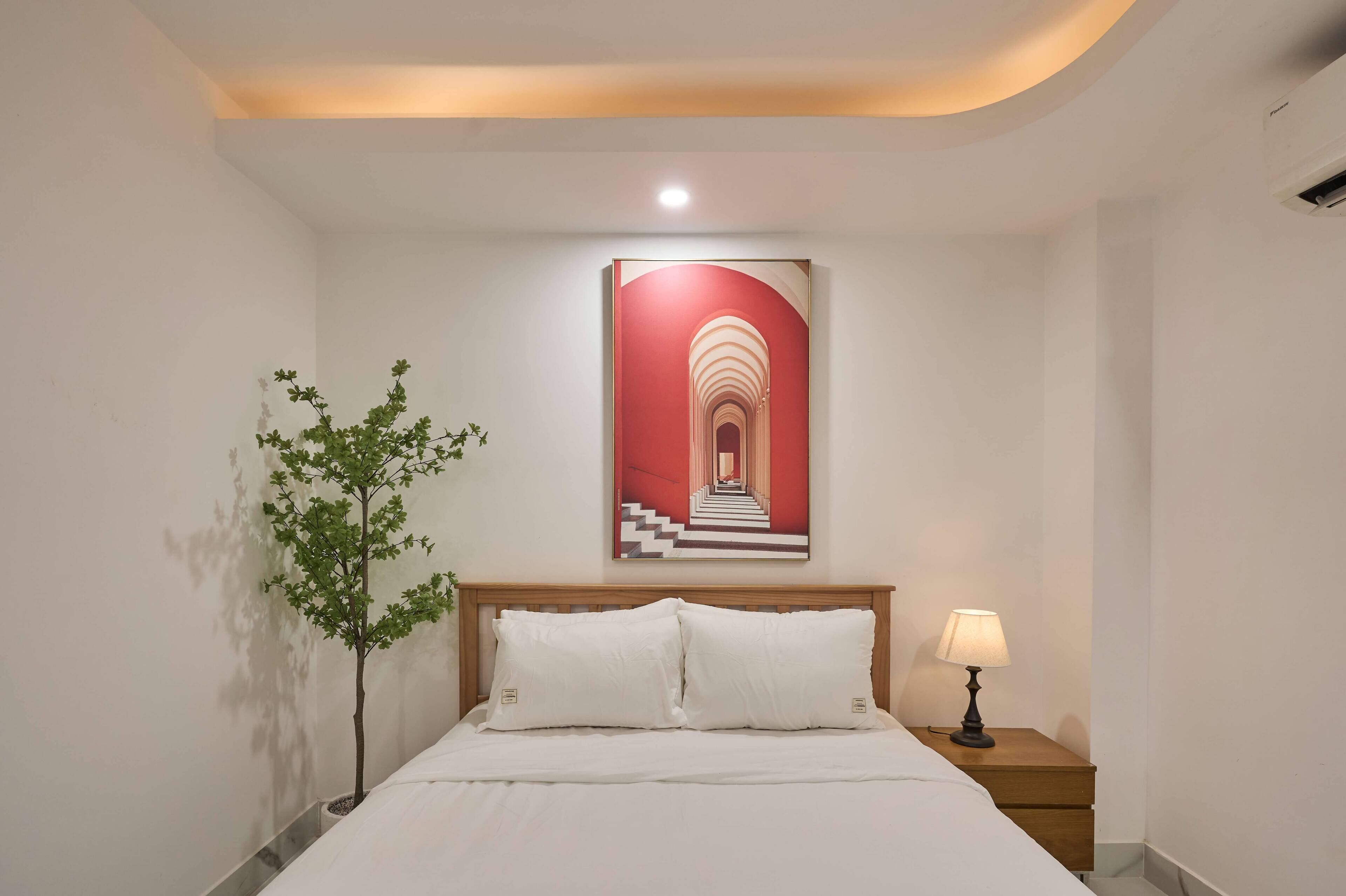 Una cama bien hecha con sábanas blancas, una mesita de noche pequeña con una lámpara y un cuadro en la pared de una escalera roja y blanca.