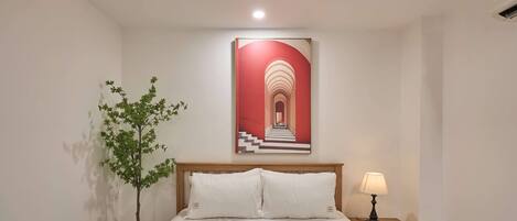 A neatly made bed with white linens, a small bedside table with a lamp, and a wall-mounted painting of a red and white staircase.