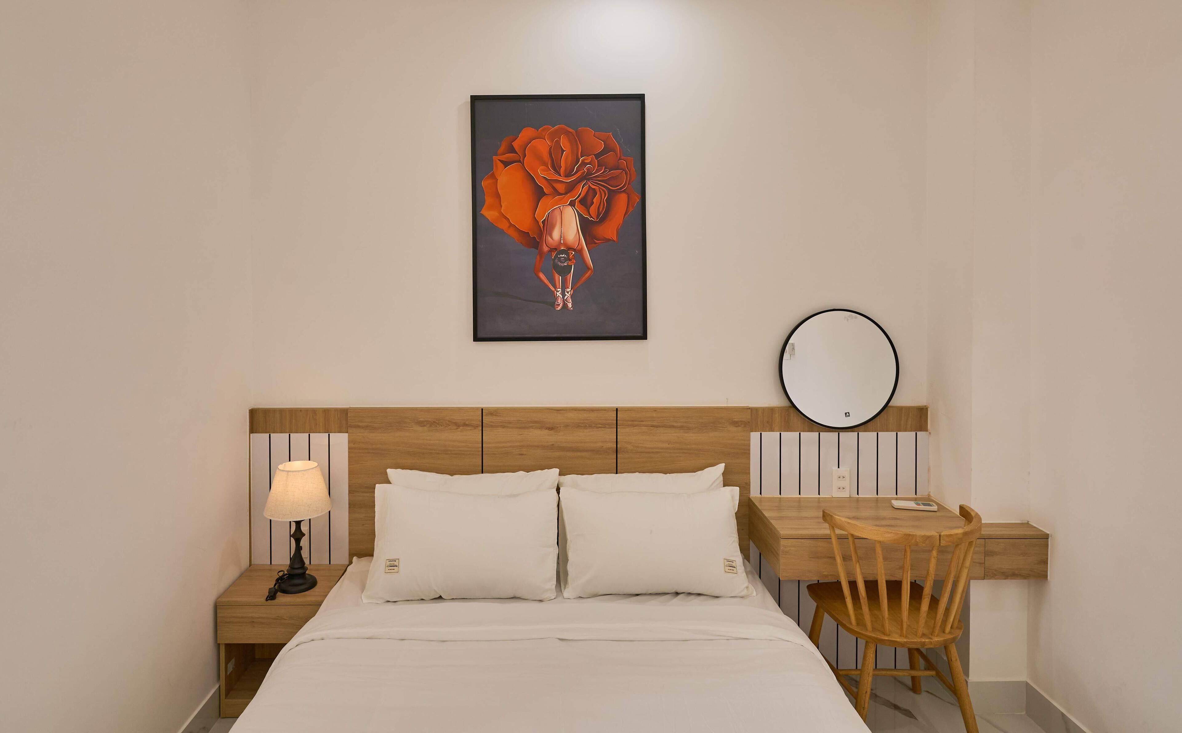 A bedroom with a bed, bedside table with a lamp, a desk with a chair, a round mirror, and a framed artwork on the wall.