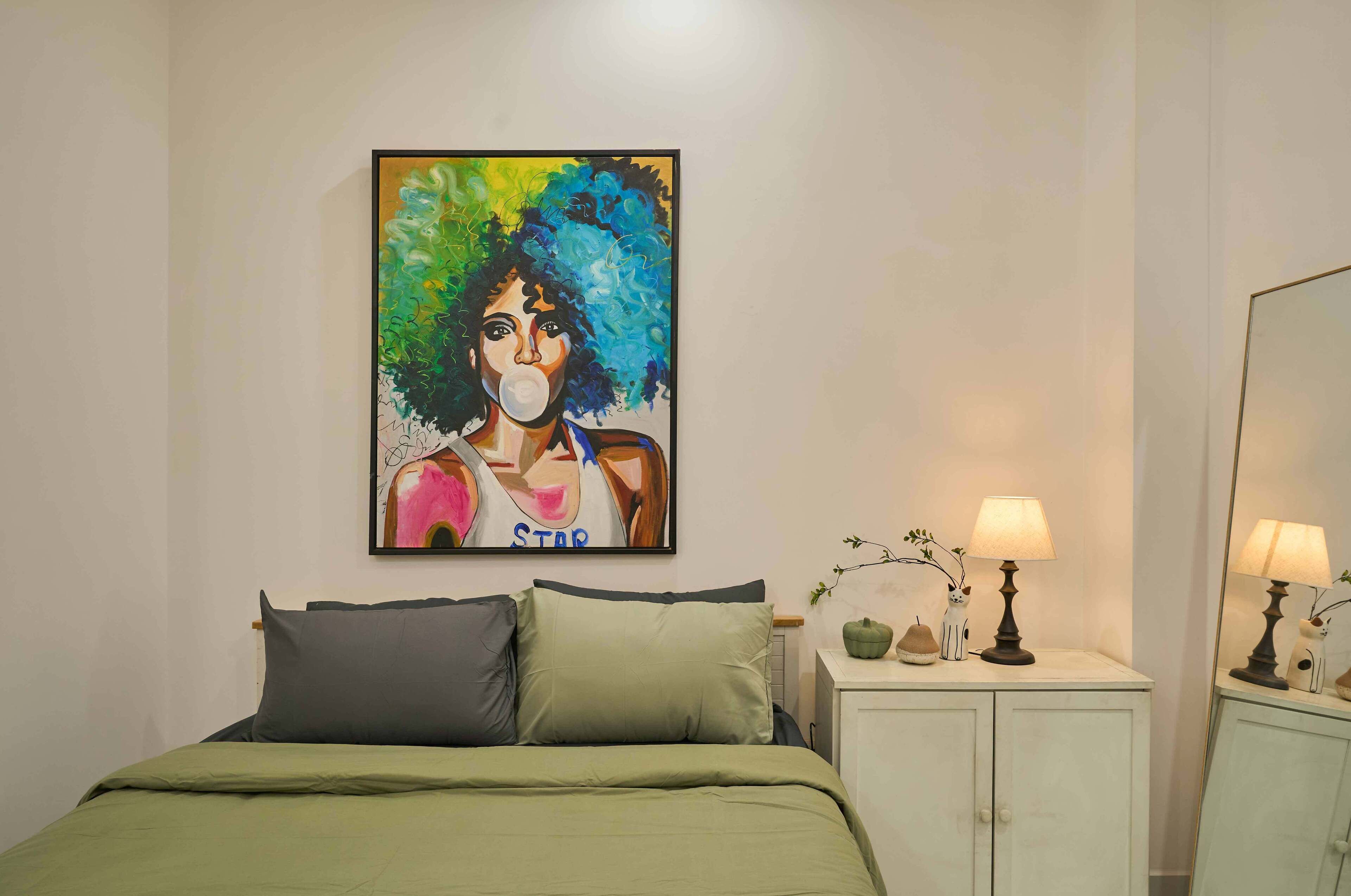 A bedroom with a bed, pillows, a nightstand with a lamp, a small table with decorative items, and a painting on the wall.