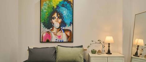 A bedroom with a bed, pillows, a nightstand with a lamp, a small table with decorative items, and a painting on the wall.