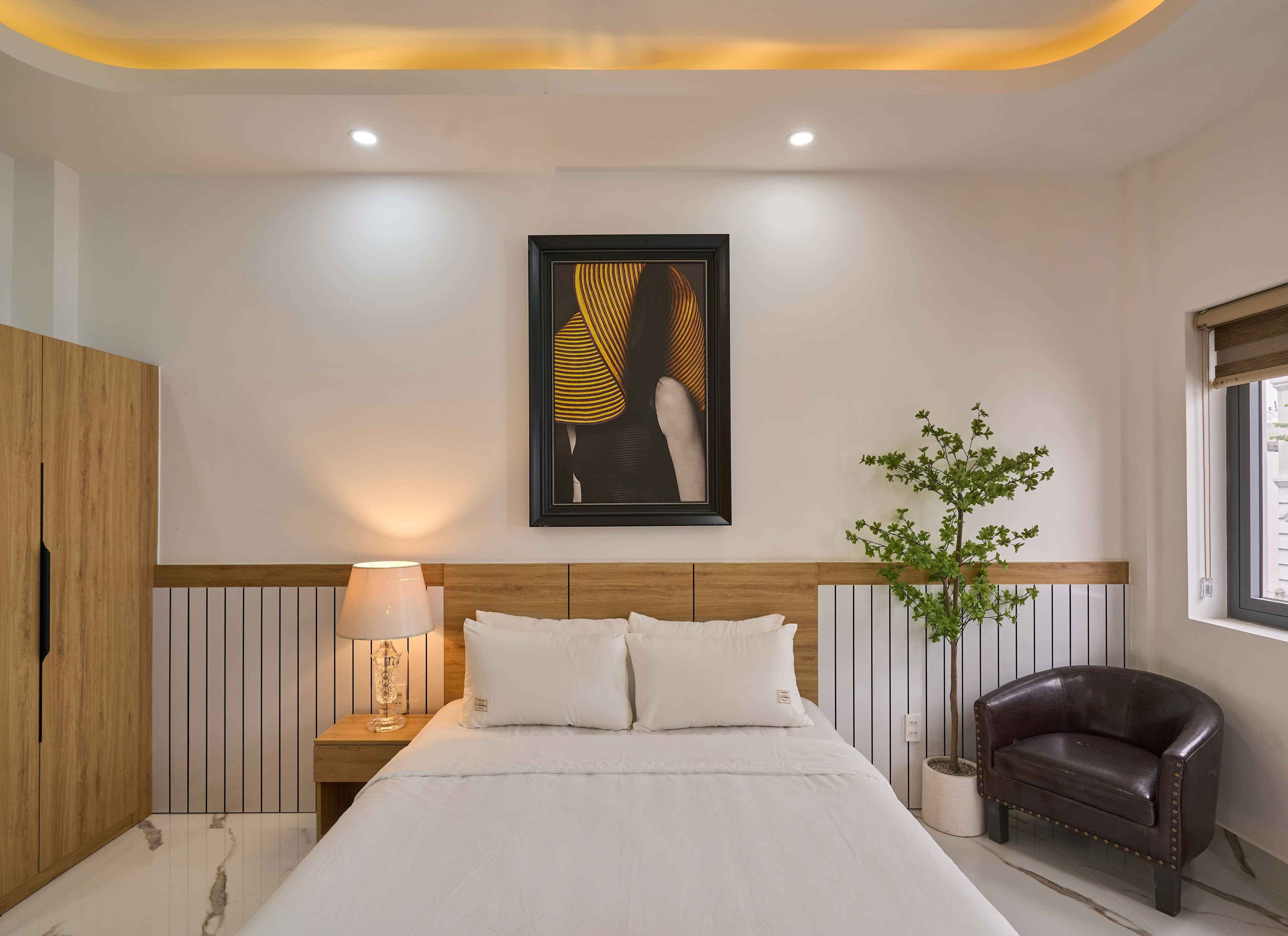Une chambre à coucher moderne avec un grand lit, une lampe de chevet, une œuvre d’art encadrée, une plante en pot et un fauteuil en cuir.
