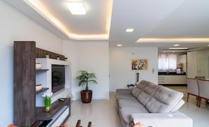 A modern living room with a grey sofa, a TV on a wooden stand, a potted plant, and a dining area in the background. - Apt w/ 3BR & BBQ 350m From the Beach Lam0601 (Itapema)