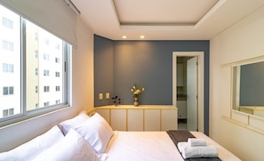 A hotel room with a bed, a desk, and a window overlooking a building. - Apt w/ 3BR & BBQ 350m From the Beach Lam0601 (Itapema)