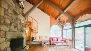 Ein rustikales Blockhaus-Interieur mit Kamin aus Stein, Holzdielen, einem roten Sofa und großen Fenstern mit Blick ins Freie.
