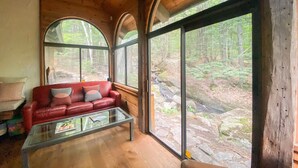 Ein gemütliches Blockhaus-Interieur mit einem roten Ledersofa, einem Glastisch und großen Fenstern mit Blick auf einen bewaldeten Bereich.