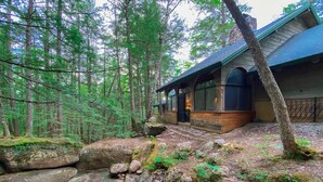 Eine rustikale Holzhütte mit Steinunterbau, großen Fenstern und einem steilen Dach, umgeben von einem bewaldeten Gebiet mit einem Bach.
