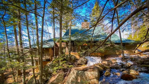 Eine rustikale Holzhütte mit einer Holzterrasse, umgeben von dichtem Wald und einem kleinen Bach.
