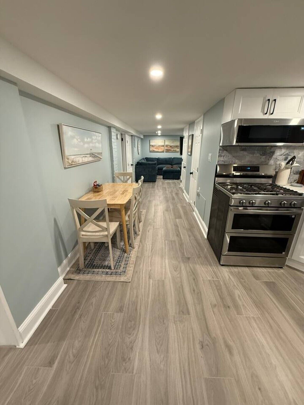 A modern kitchen with a built-in oven and microwave, a dining area with a wooden table and chairs, and a hallway leading to a living area with a sofa.