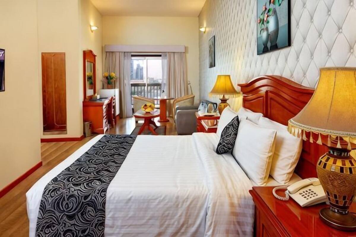 A hotel room with a bed, bedside table, lamp, and a view of the city.