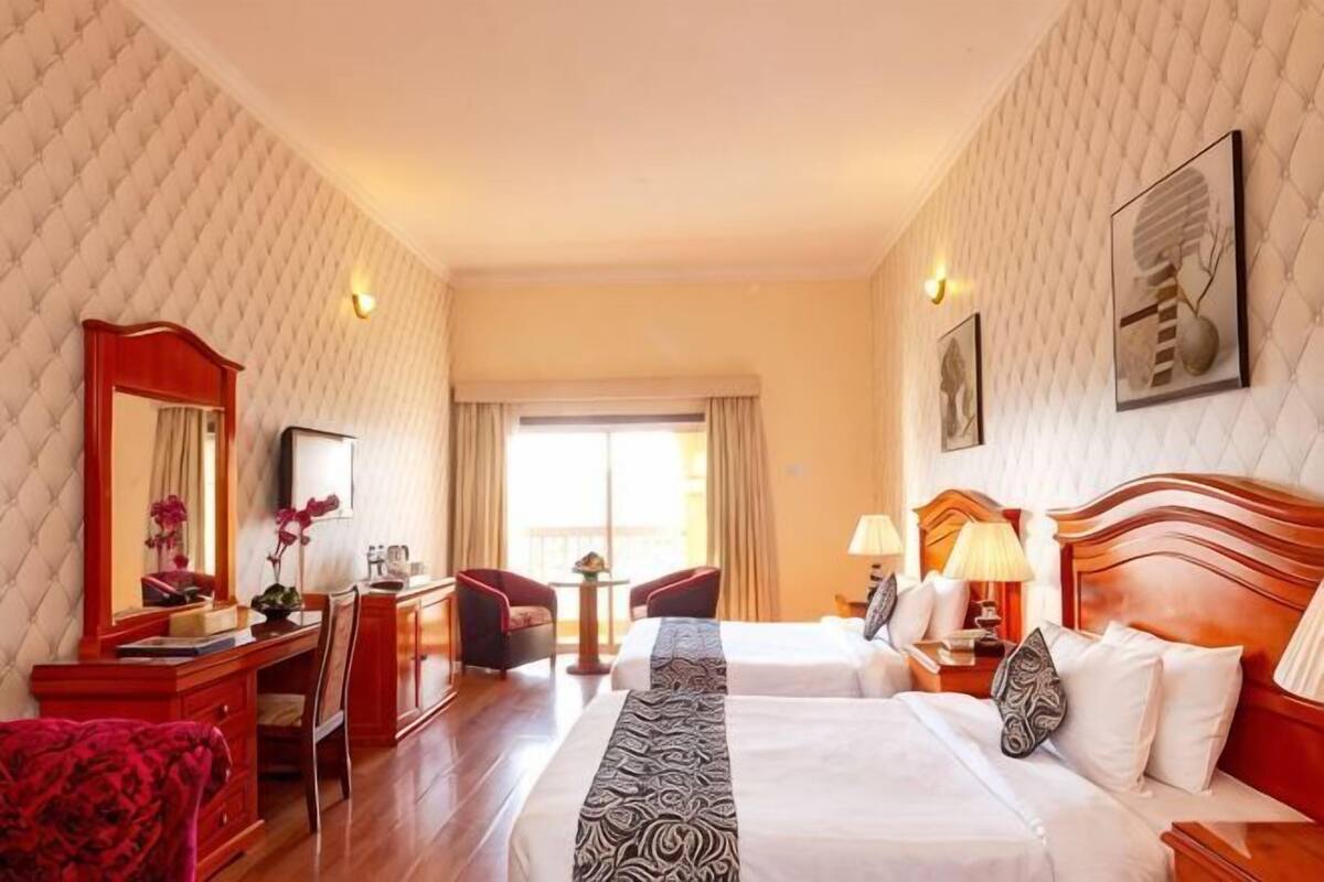 A hotel room with two beds, a desk, a chair, a red sofa, a mirror, and a window with curtains.