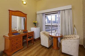 A hotel room with a wooden dresser, a desk, two armchairs, a small table, a potted plant, and a large window with curtains. - Gulf Gate Hotel (Manama)