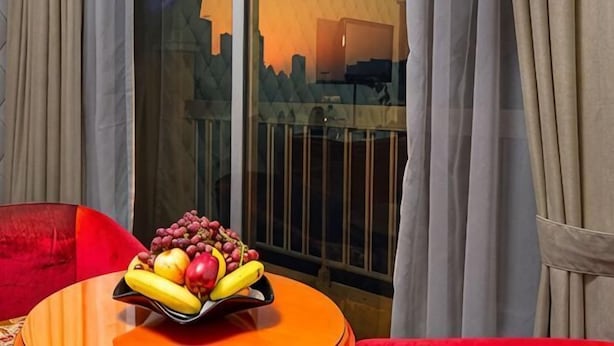 A room with a table, a bowl of fruit, and a view through a window with curtains. - Gulf Gate Hotel (Manama)