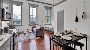 Un apartamento moderno con cocina, comedor y sala de estar. Cuenta con ventanas amplias, un sofá gris y una mesa de comedor preparada para cuatro personas.