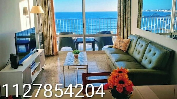 A living room with a television, a sofa, a coffee table, and a view of the sea through large windows. - Charming Mar del Plata Apartment With Ocean Views (Mar del Plata)