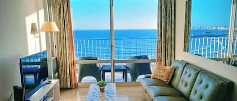 A living room with a television, a sofa, a coffee table, and a view of the sea through large windows.