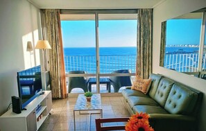 A living room with a television, a sofa, a coffee table, and a view of the sea through large windows. - Charming Mar del Plata Apartment With Ocean Views (Mar del Plata)