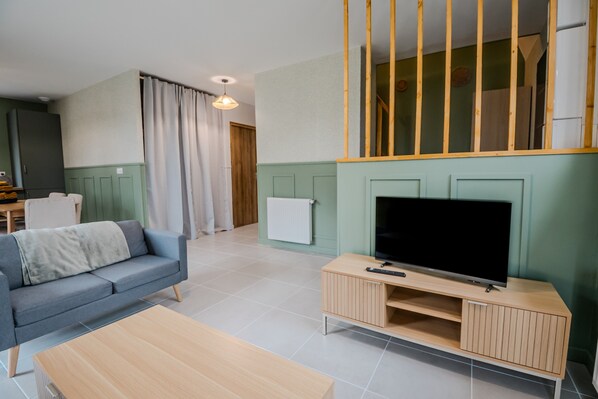 A modern living room with a sofa, a coffee table, a TV on a wooden stand, and a dining area in the background. - Gîte "Gîte Du Pré Simon" with Private Garden and Wi-Fi (Le Faulq)
