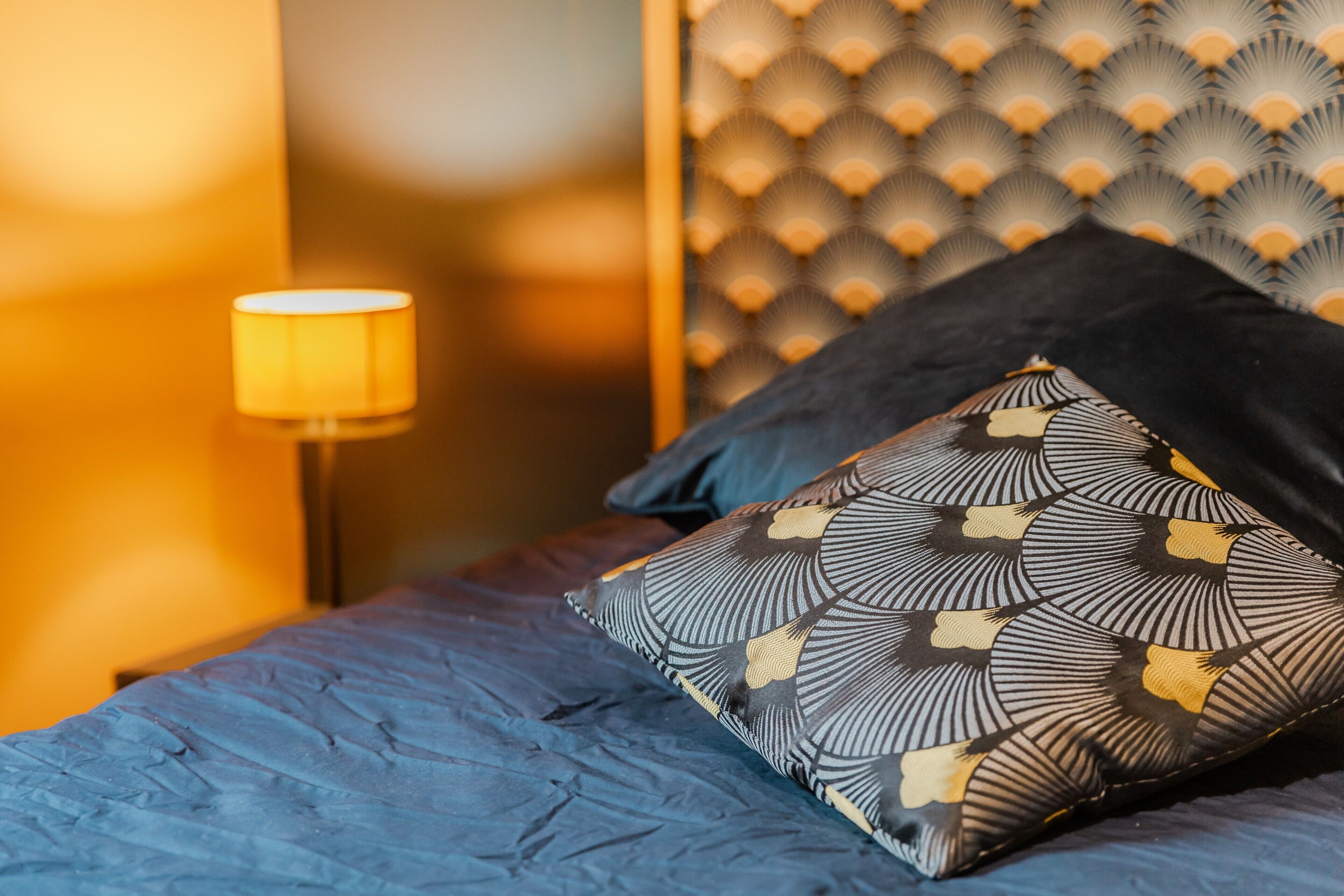 Una cama con un almohadón estampado, un cabecero de diseño geométrico y una lámpara sobre una mesita de noche.