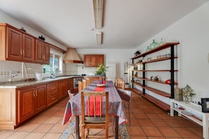 A kitchen with wooden cabinets, a dining table with chairs, a tiled floor, and a shelf with decorative items.