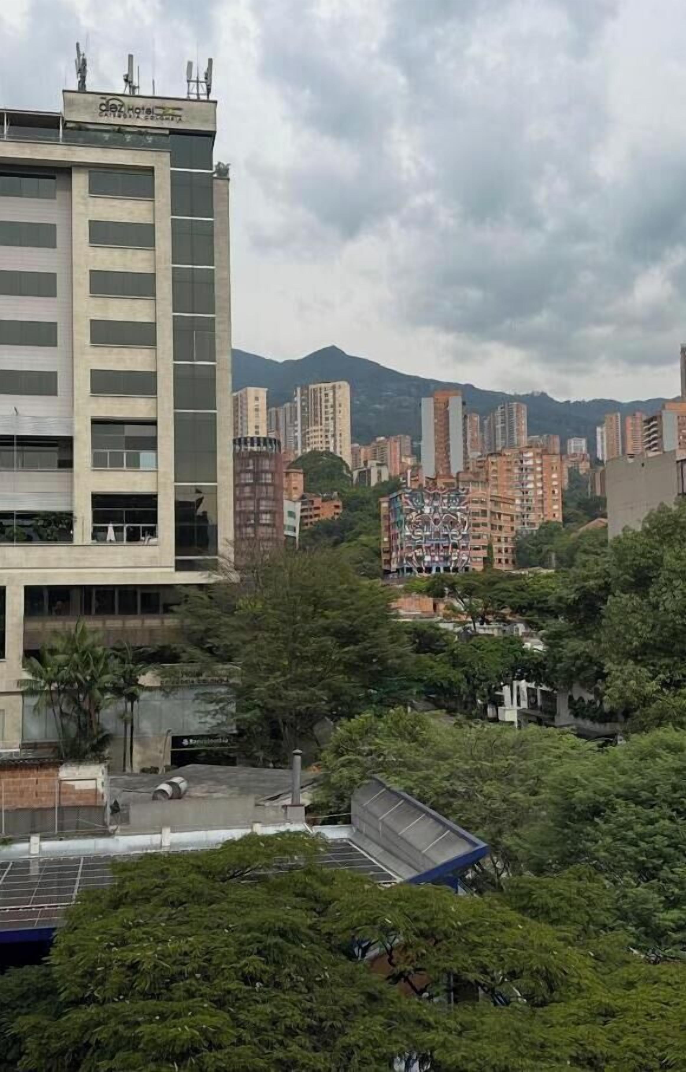 Un hotel de diverses plantes amb façana plana, envoltat de vegetació abundant i altres edificis al fons.
