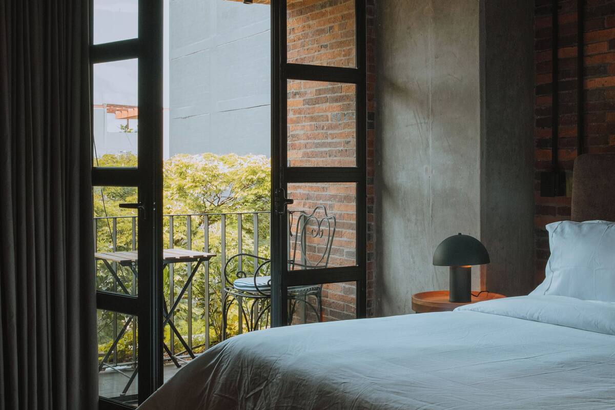 Un dormitorio con una cama grande, una mesita de noche, una lámpara y vistas a un balcón con sillas.