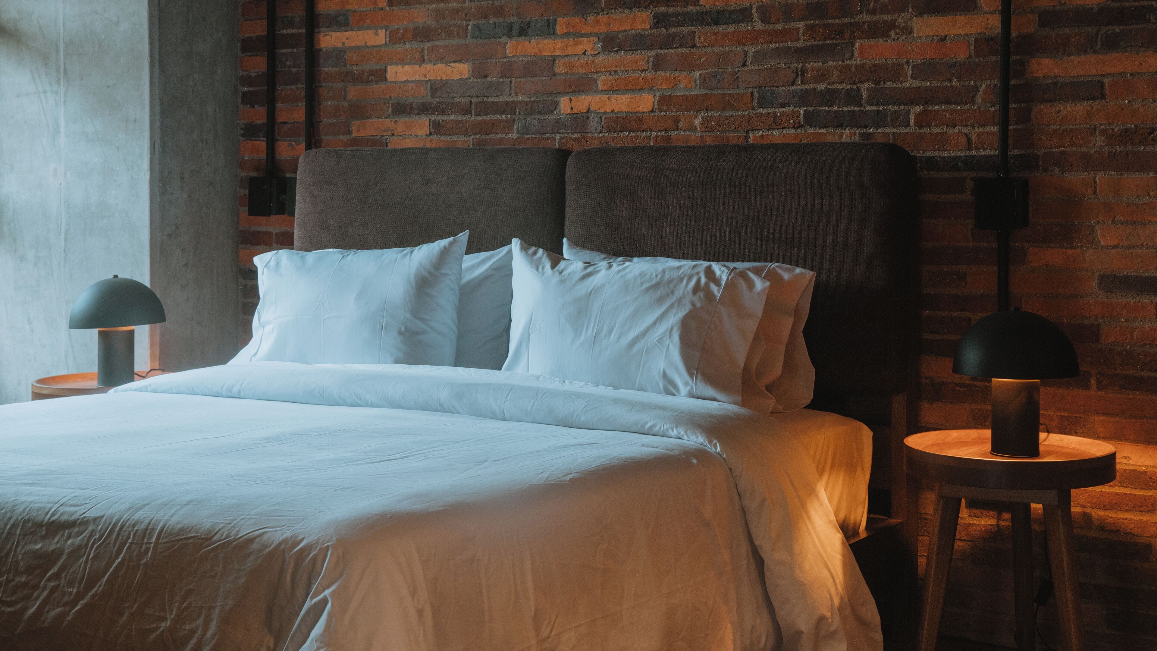 Une chambre moderne avec un mur en briques, un grand lit recouvert de draps blancs et deux lampes de chevet.