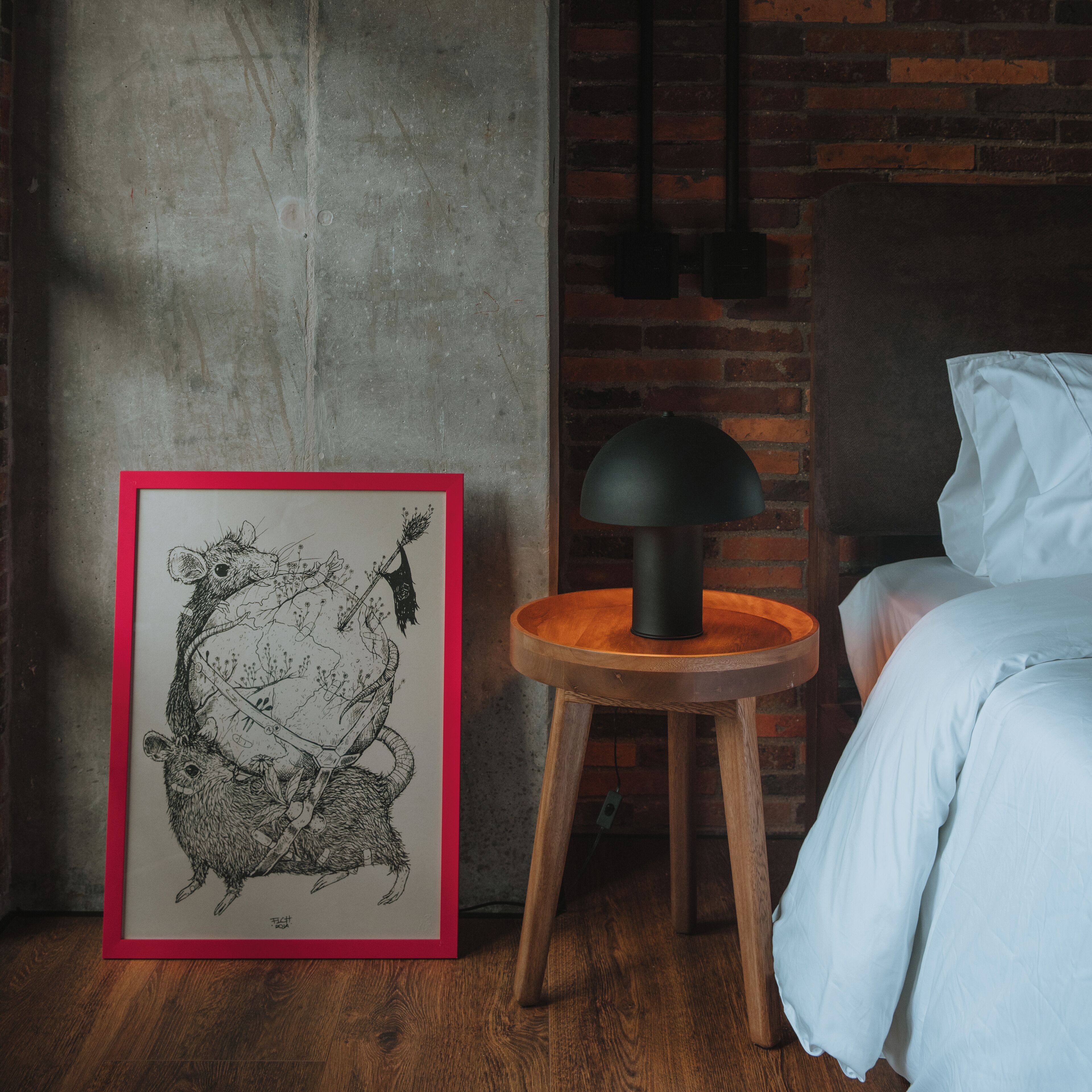 A framed illustration of a bear on a wall, a round wooden table with a black lamp, and a bed with white bedding.