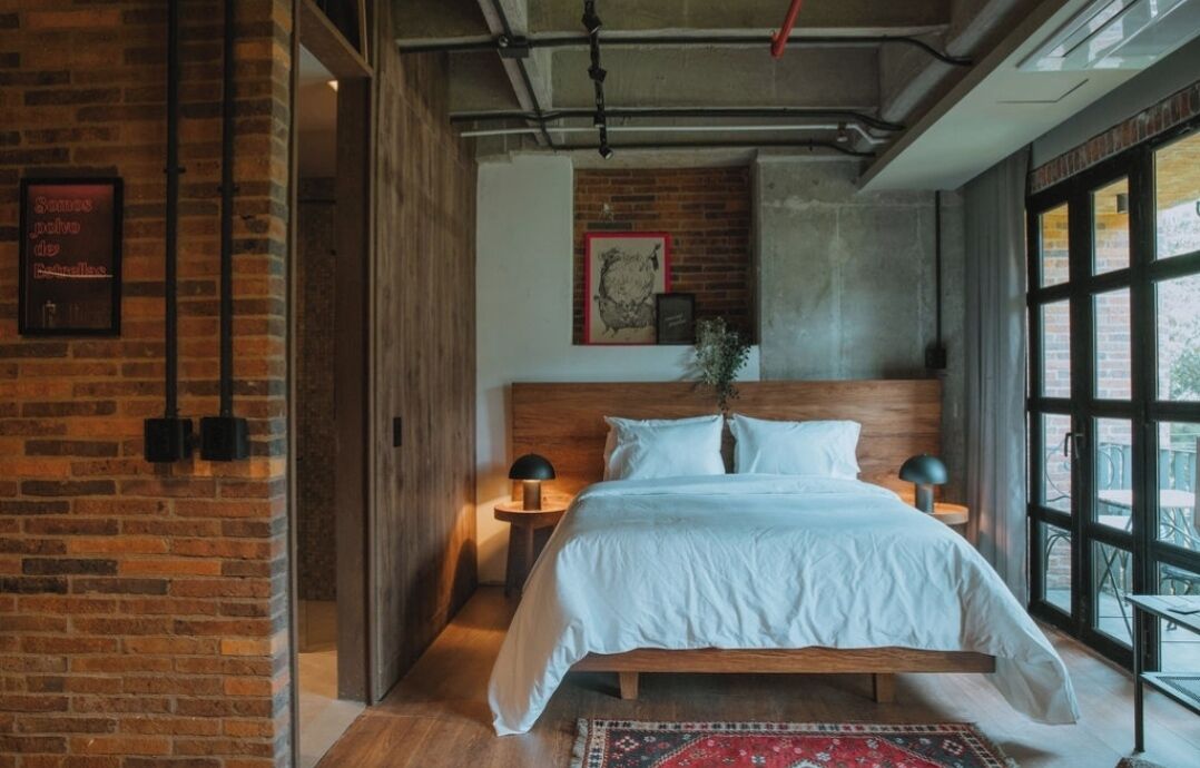 A bedroom with a wooden bed, a brick wall, a large window, and a small side table with a lamp.