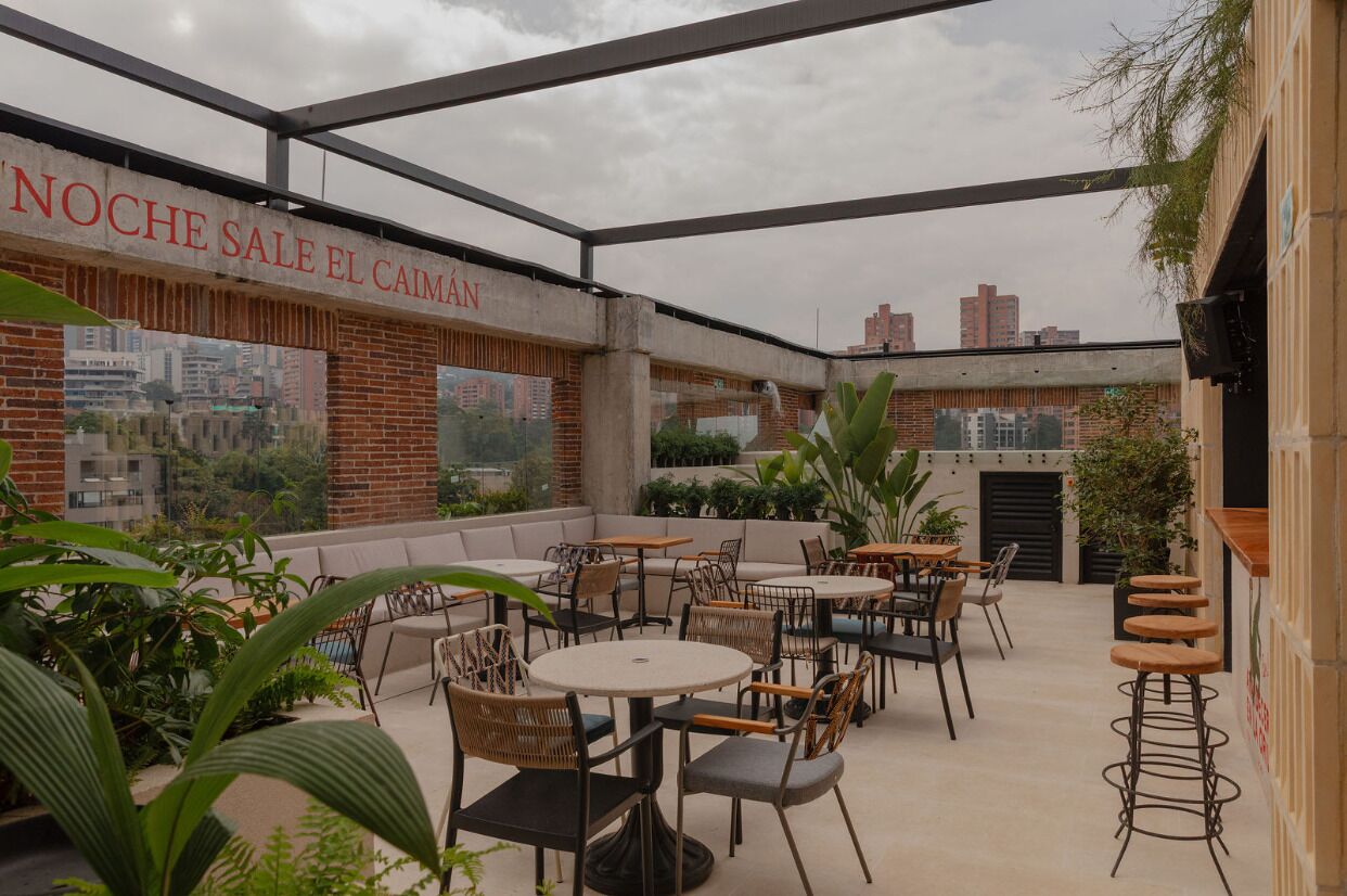 An outdoor seating area with tables and chairs, a brick wall with a mural, and a sign reading 'Noche Sale El Caiman'.