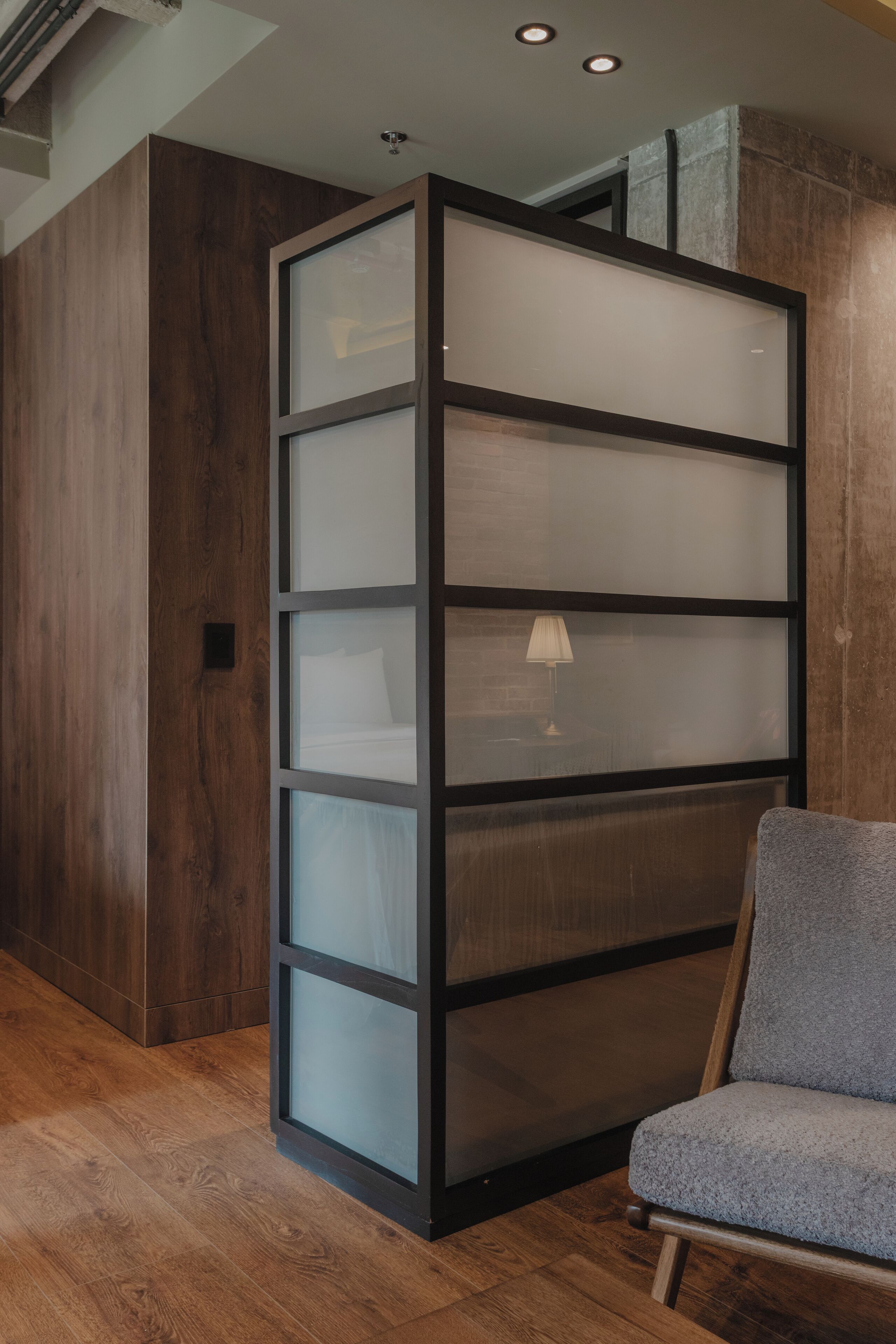 Una habitación con piso de madera, un sillón tapizado en gris y un separador alto y estrecho con paneles de vidrio esmerilado.